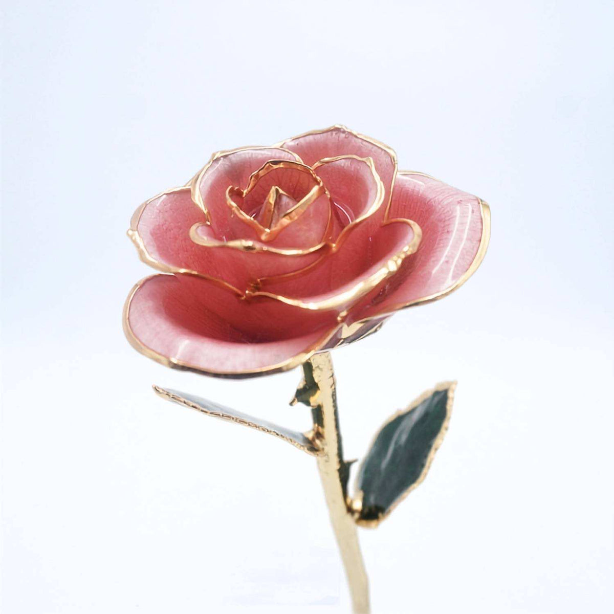A single decorative rose with pink enamel petals outlined in gold, attached to a gold stem with a dark green leaf, photographed against a clean white background.